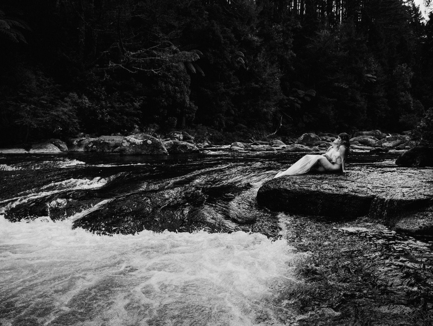 Black and white photo of a person reclining on rocks beside a rushing river surrounded by dark forest.