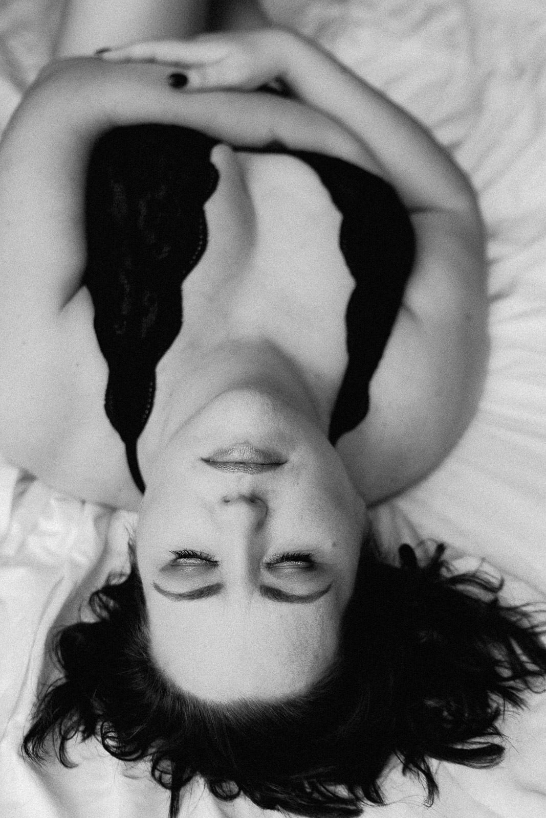 Black and white boudoir photo of a woman lying on white bedding wearing black lace lingerie.