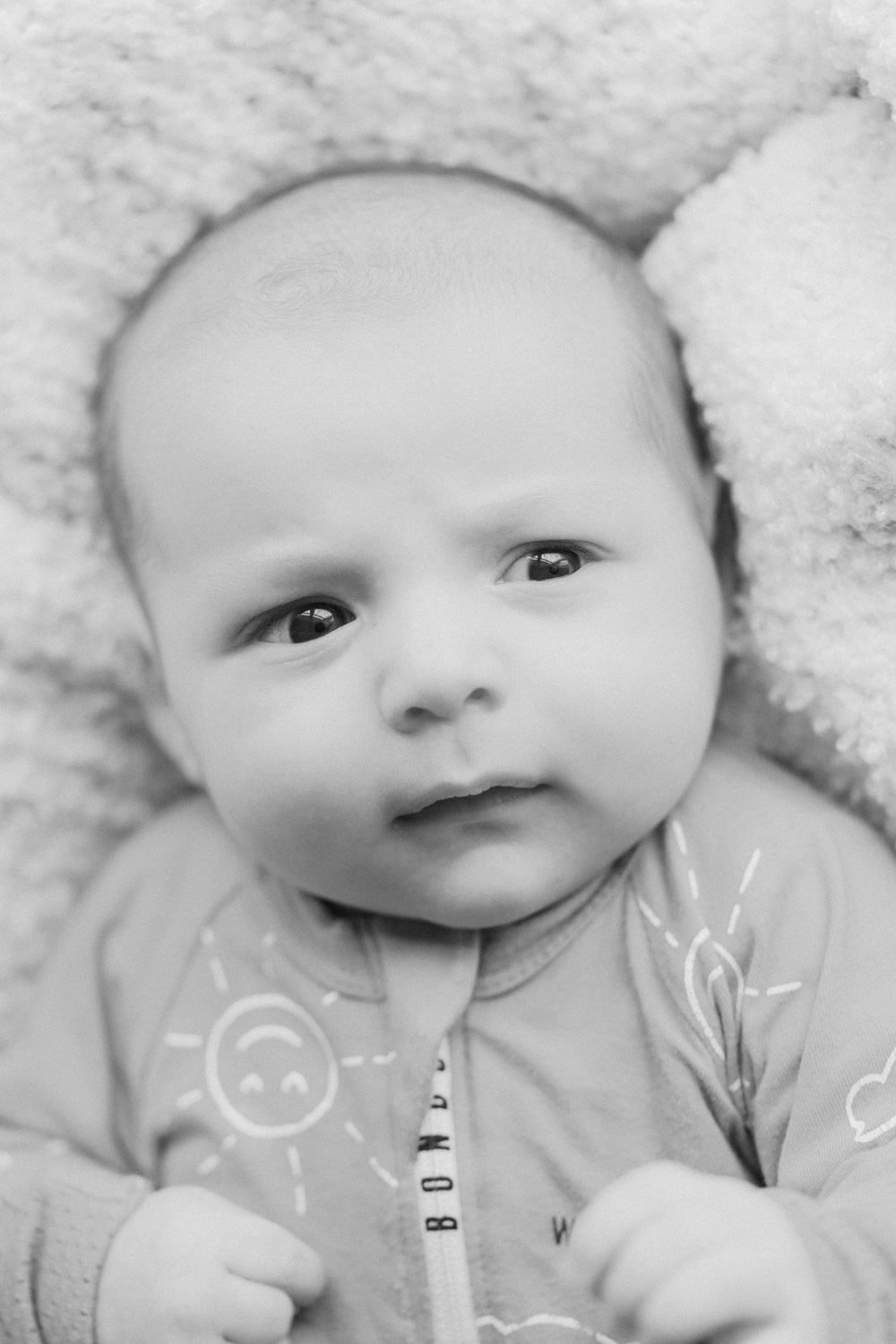 Black and white portrait of an alert newborn baby wearing a sun-print onesie, lying on a soft fluffy blanket.