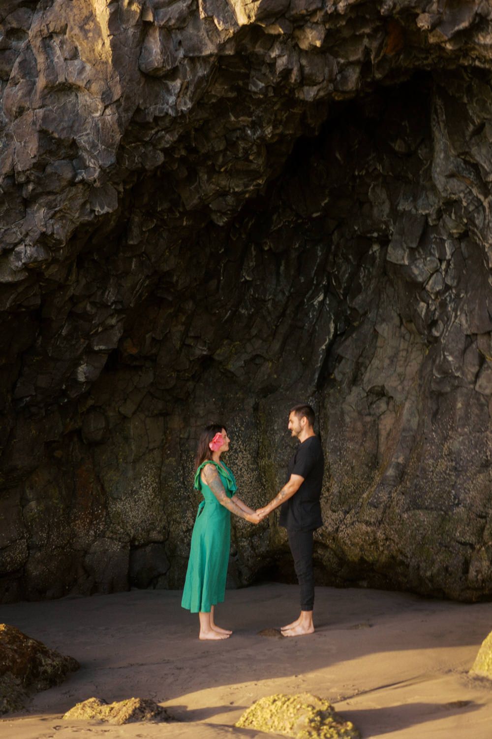 Couple holding hands in front of a dramatic rocky cave on a sandy beach, woman in green dress, man in black outfit.