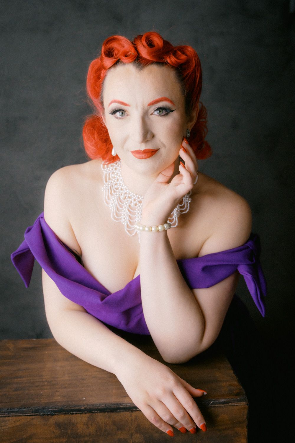 A glamorous woman with red pin-up hair wearing a purple off-shoulder dress and pearl jewelry poses elegantly.
