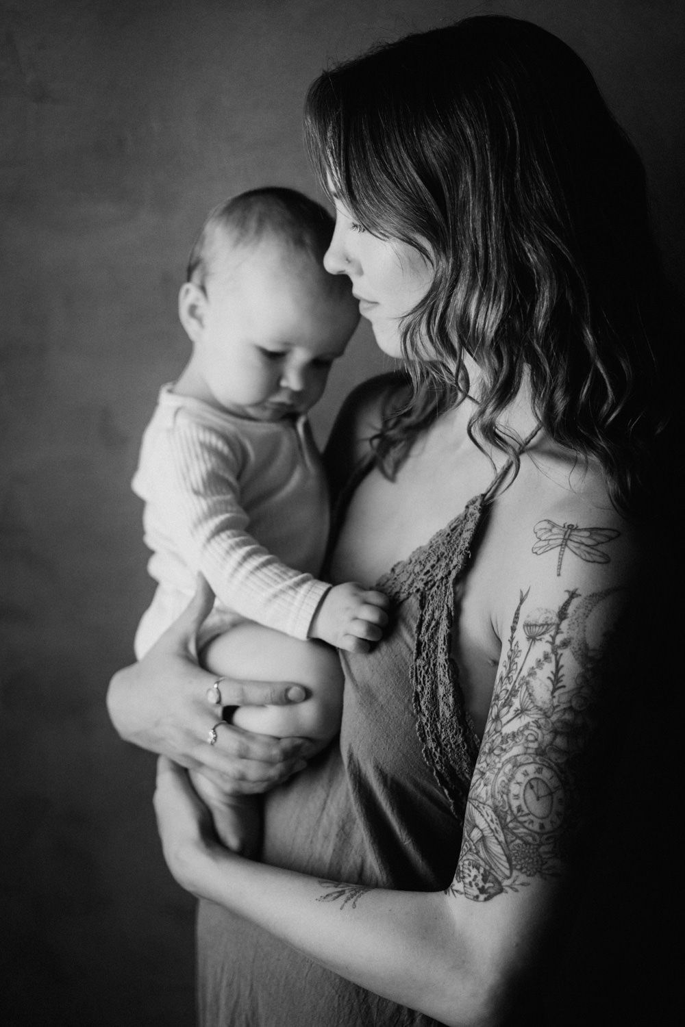 Black and white portrait of a tattooed mother tenderly holding her baby, captured in soft studio lighting.