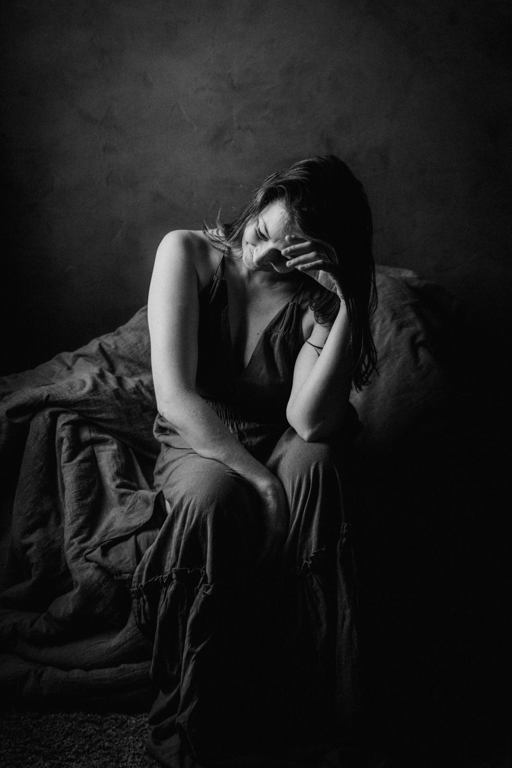 Black and white photo of a sad woman sitting on a bed with her face in her hand in a dark room.