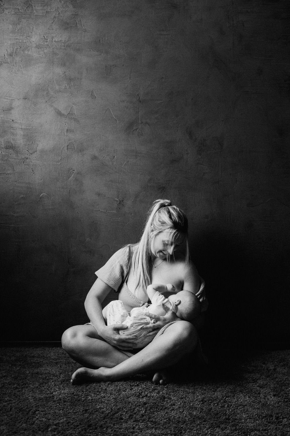 Black and white photo of a mother breastfeeding her newborn baby while sitting cross-legged on the floor.