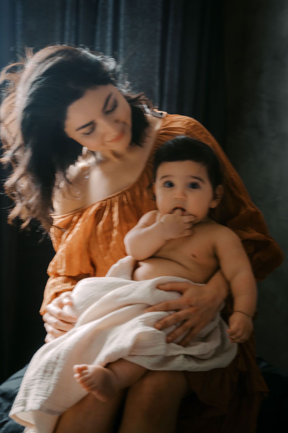 A smiling woman in an orange dress tenderly holds a baby wrapped in a white blanket on her lap.