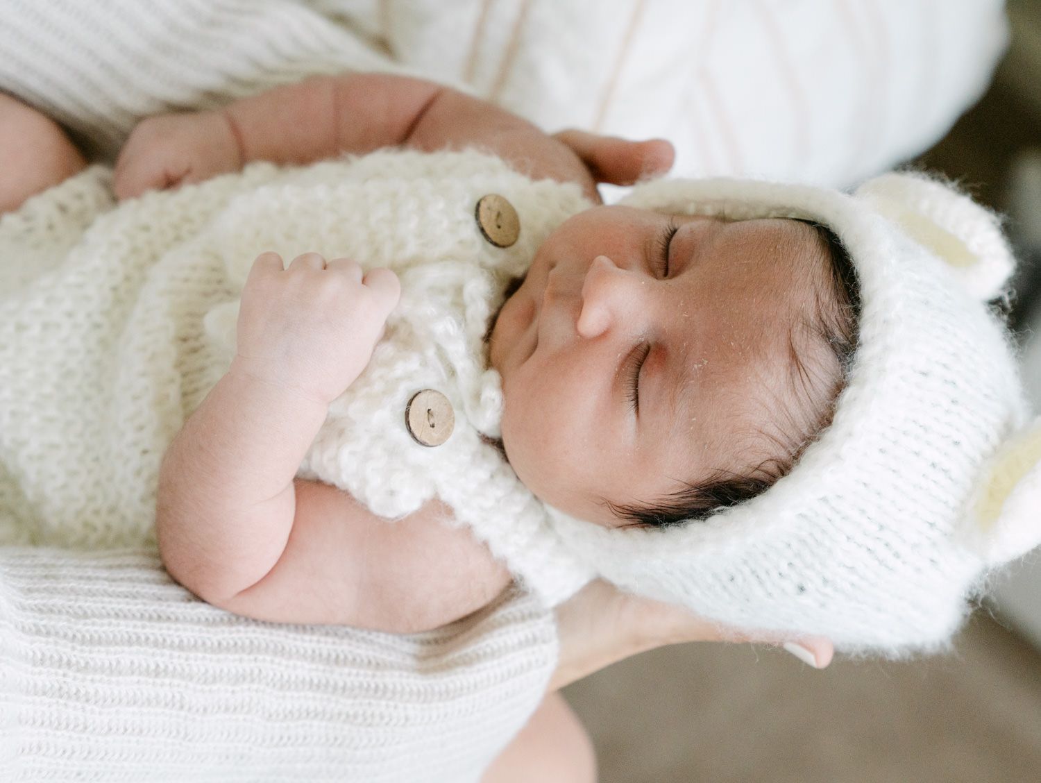 Sleeping newborn baby in white knit bear bonnet and button cardigan cradled in arms.