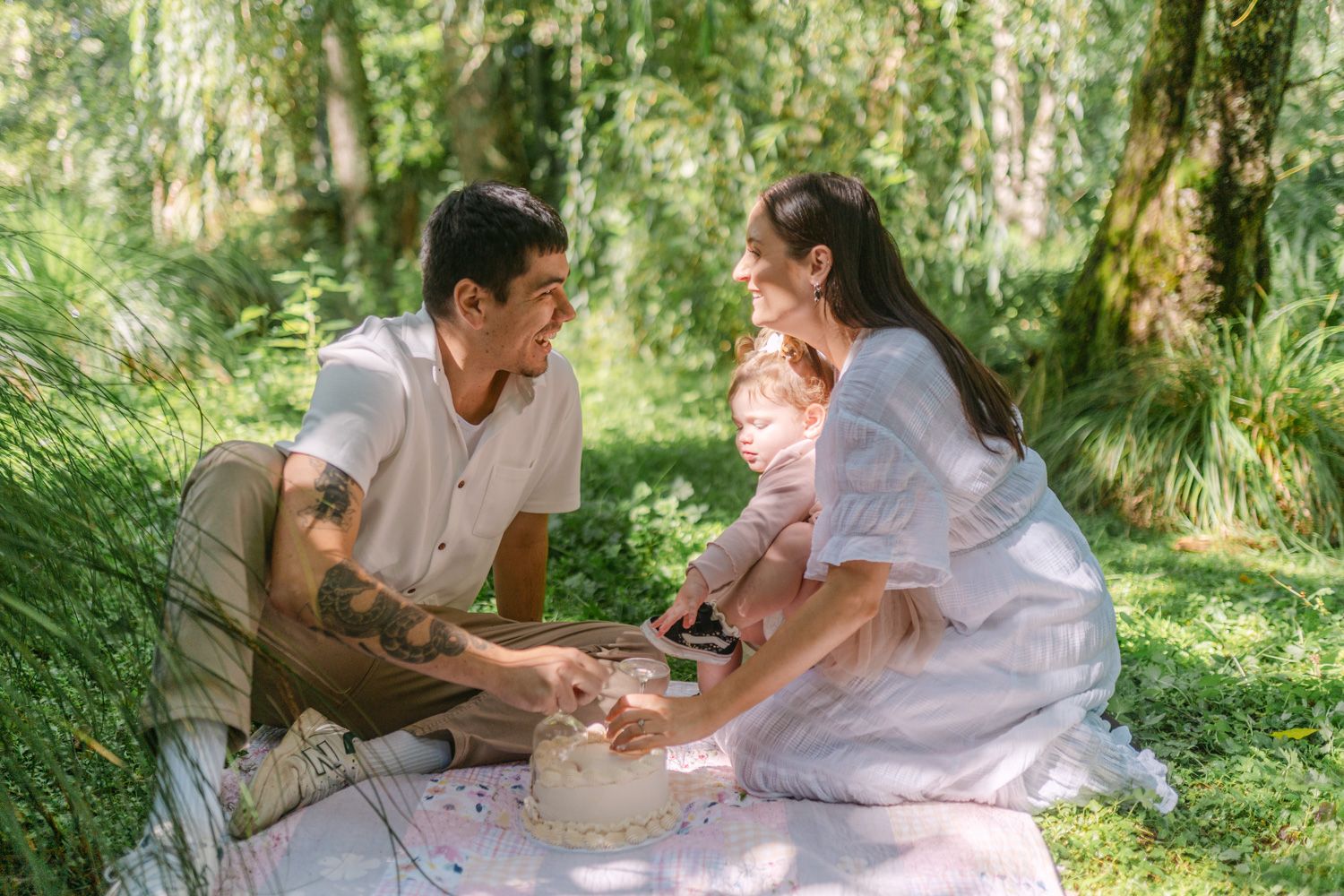 Young family enjoying a sunny outdoor picnic on a blanket in a lush green forest with their baby.
