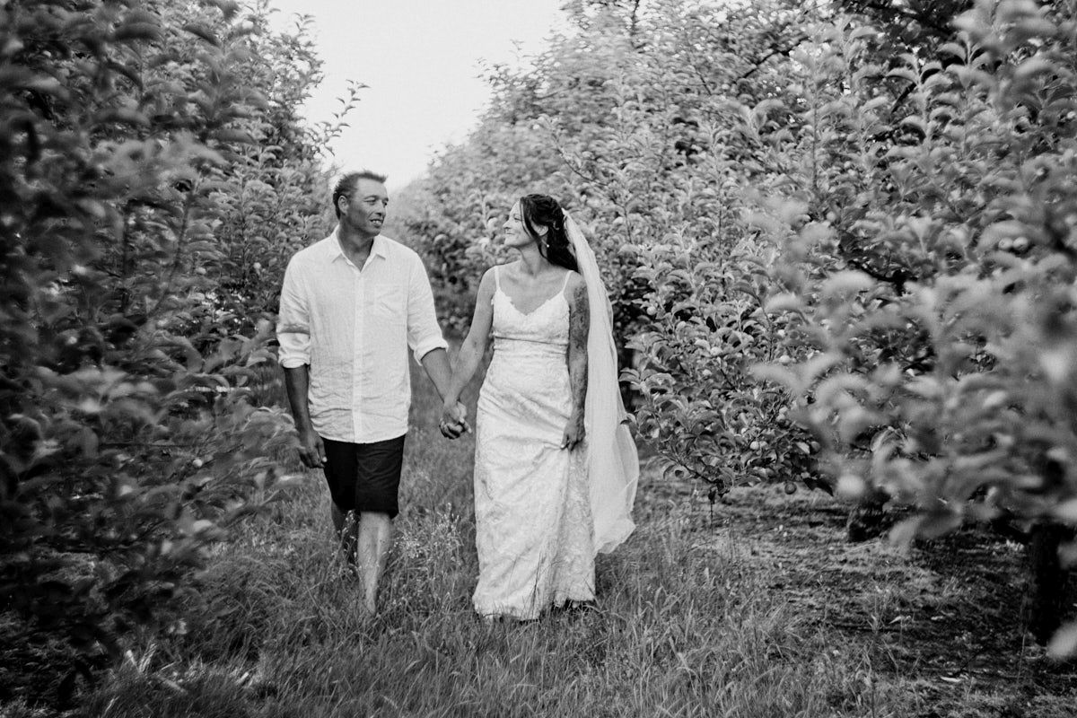 Black and white photo of a bride and groom holding hands walking through a lush orchard on their wedding day.