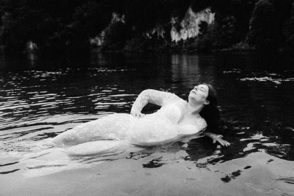 Black and white photo of a woman in a lace dress floating gracefully in a calm natural lake.
