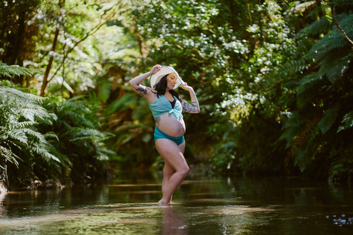 Pregnant woman in teal bikini and hat poses gracefully in a lush green forest stream surrounded by ferns.