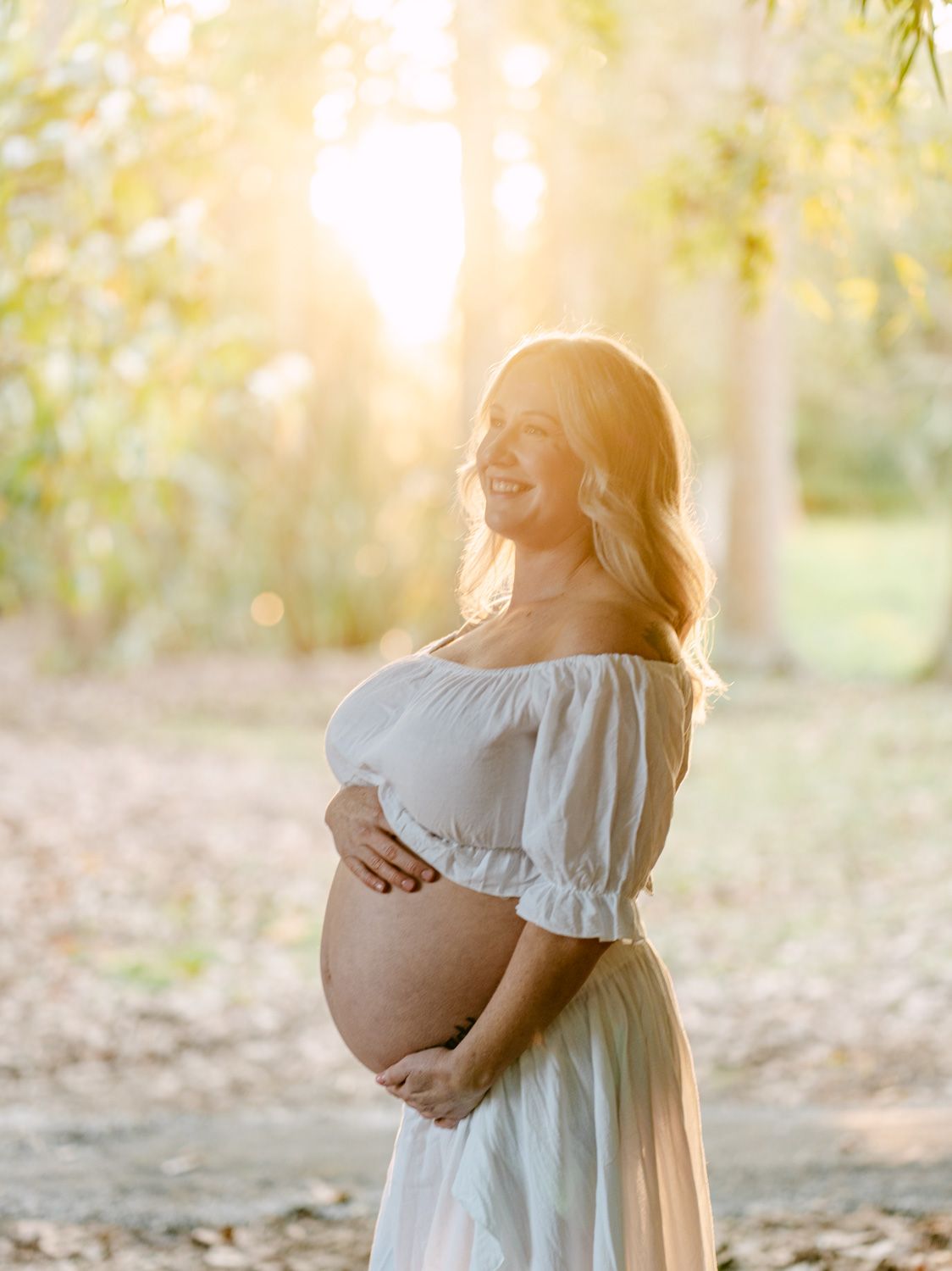 Smiling pregnant woman in white off-shoulder dress cradling baby bump outdoors in golden sunlight among trees.