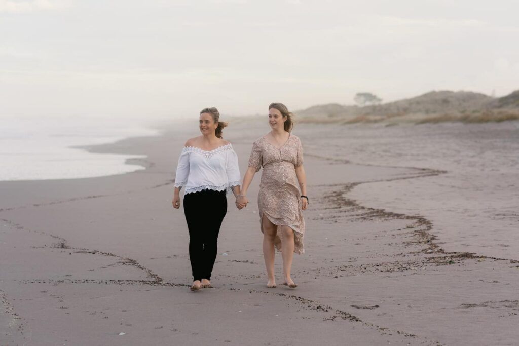 Two women walking hand in hand along a serene beach at dusk, one in white top and black pants, other in beige floral dress.
