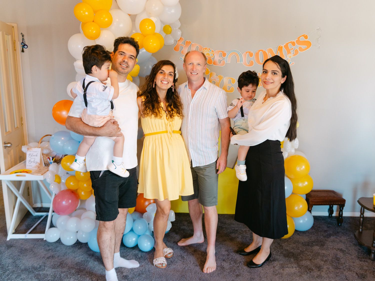 Family group photo at a festive baby shower with colorful balloon decorations in yellow, white, and blue.