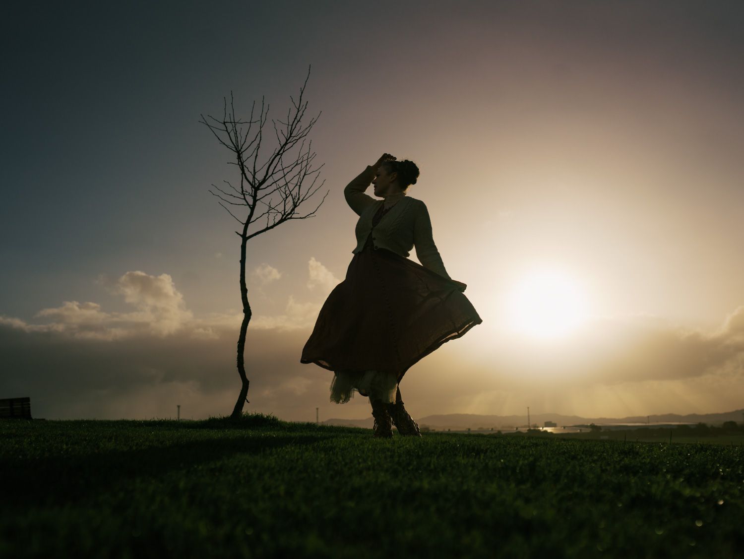Silhouette of a woman in a flowing dress and hat dancing near a bare tree at sunset.