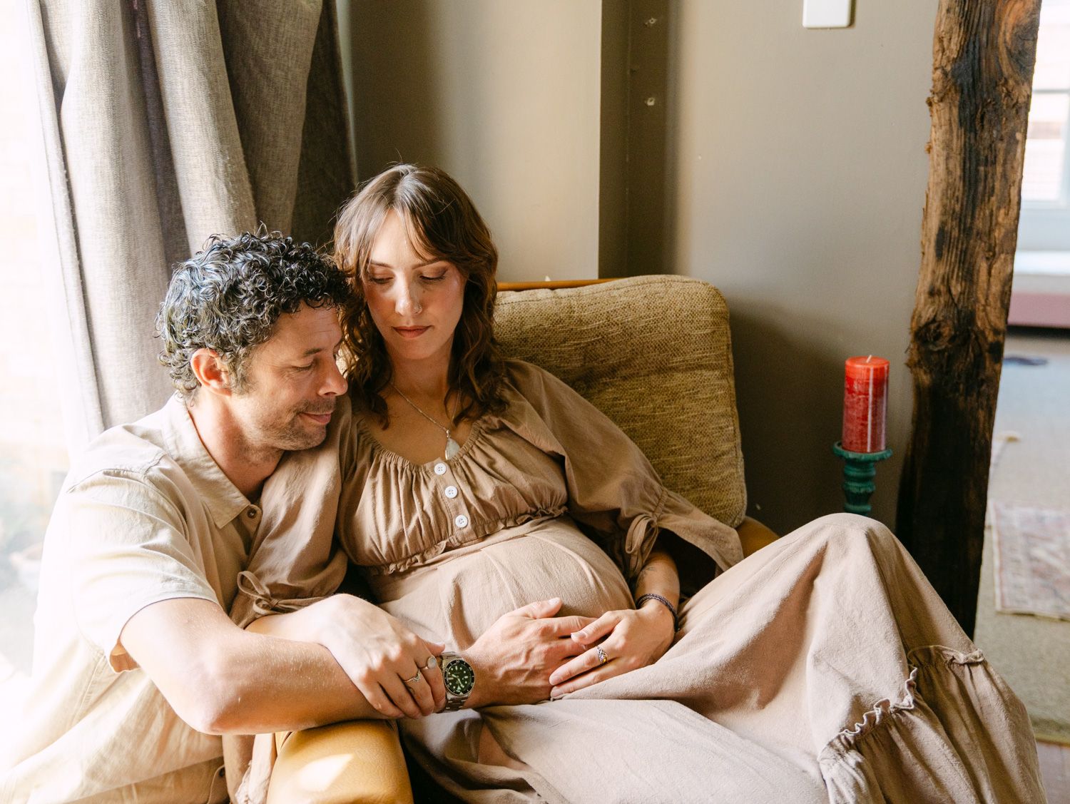 A couple tenderly cradles a pregnant belly while sitting together in a cozy armchair by a sunlit window.