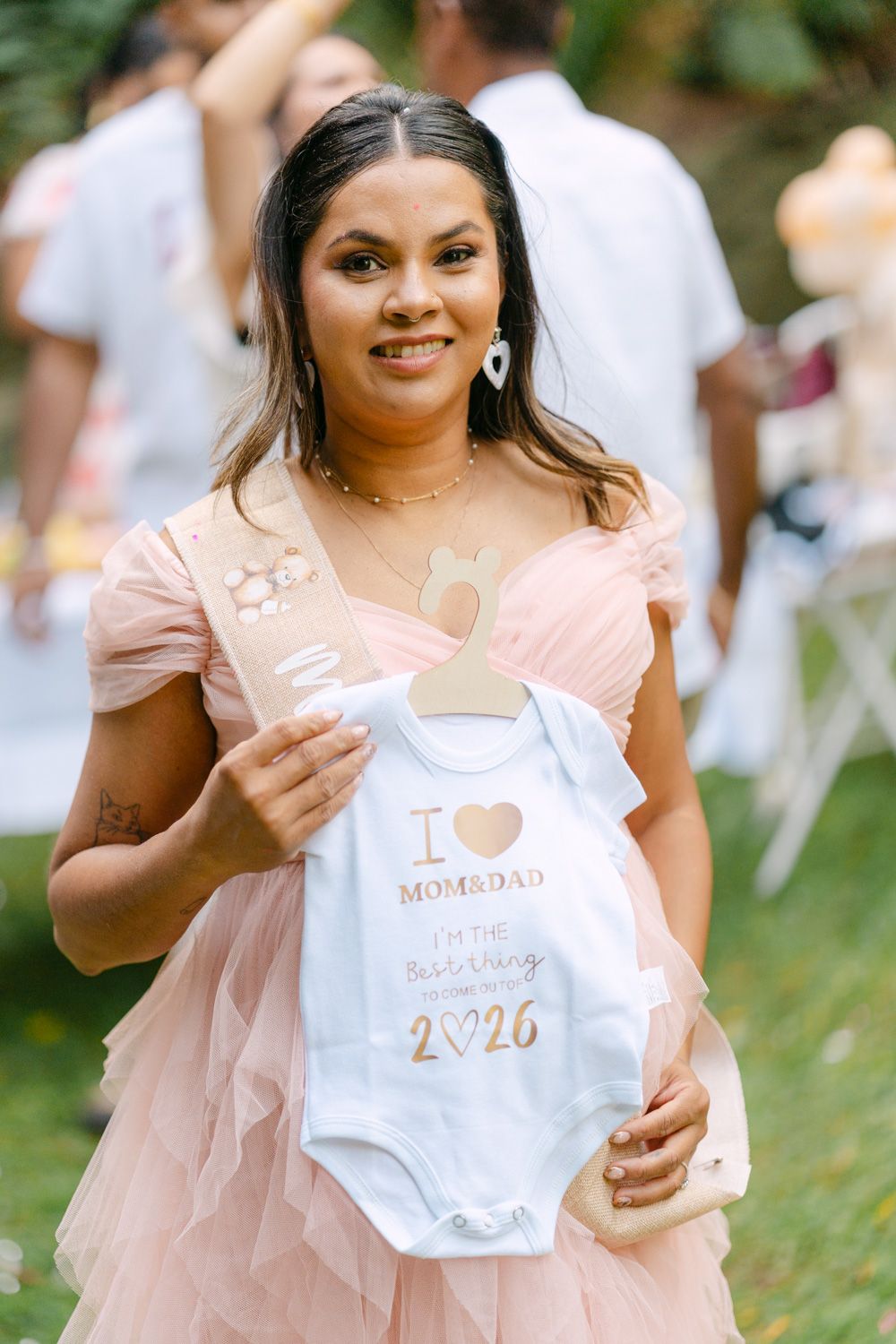 Woman in pink tulle dress holding a baby onesie reading I Love Mom & Dad at an outdoor baby shower.