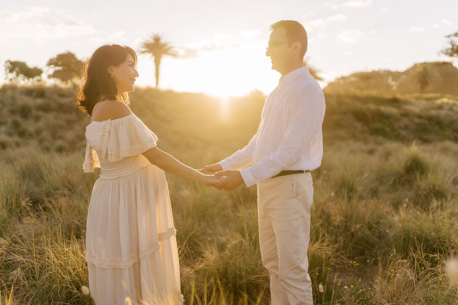 Couple holding hands in a golden sunset field, woman in white off-shoulder dress, man in white shirt and khaki pants.