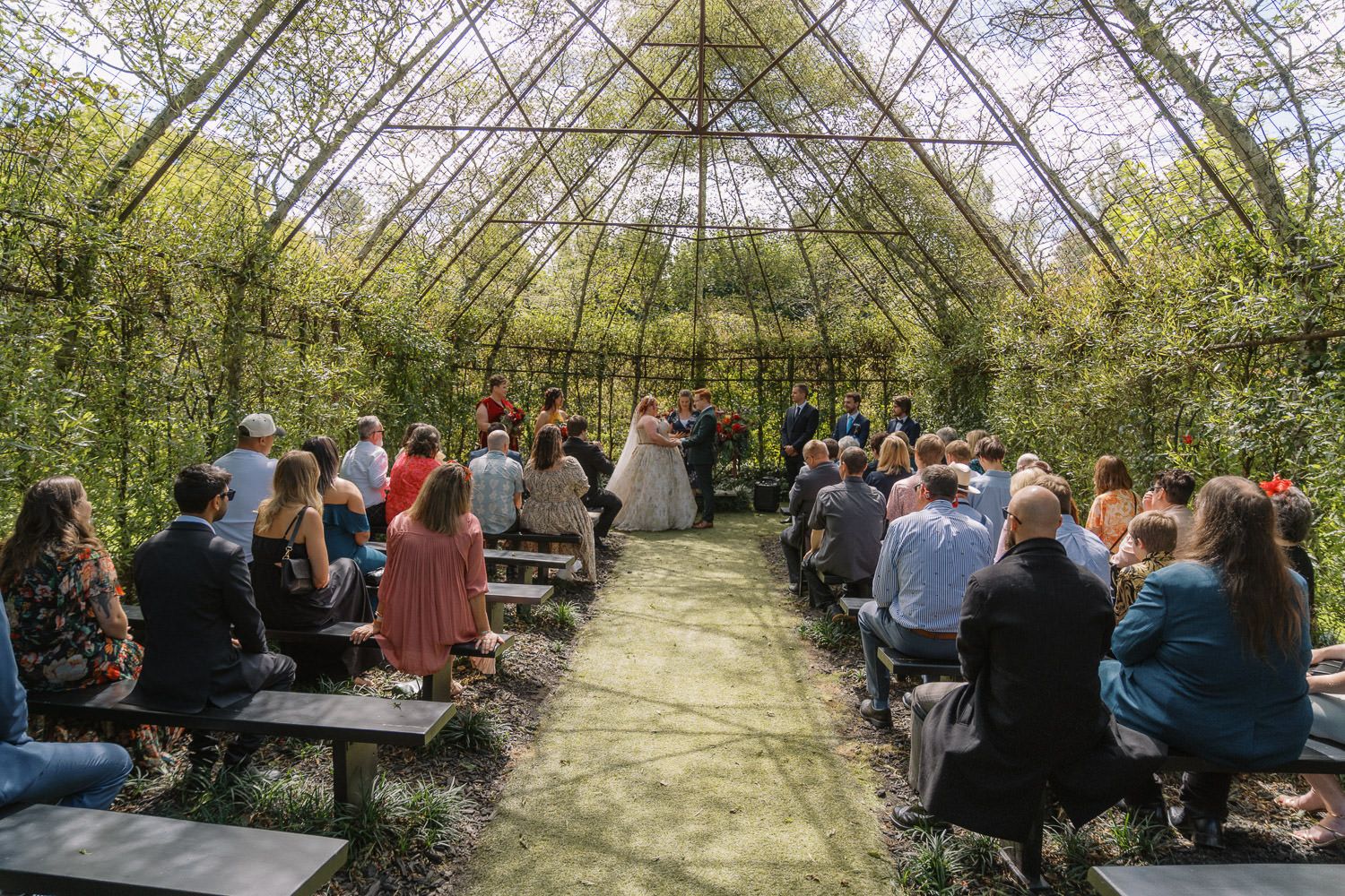 Outdoor wedding ceremony in a lush green botanical garden pavilion with guests seated on wooden benches facing the altar.