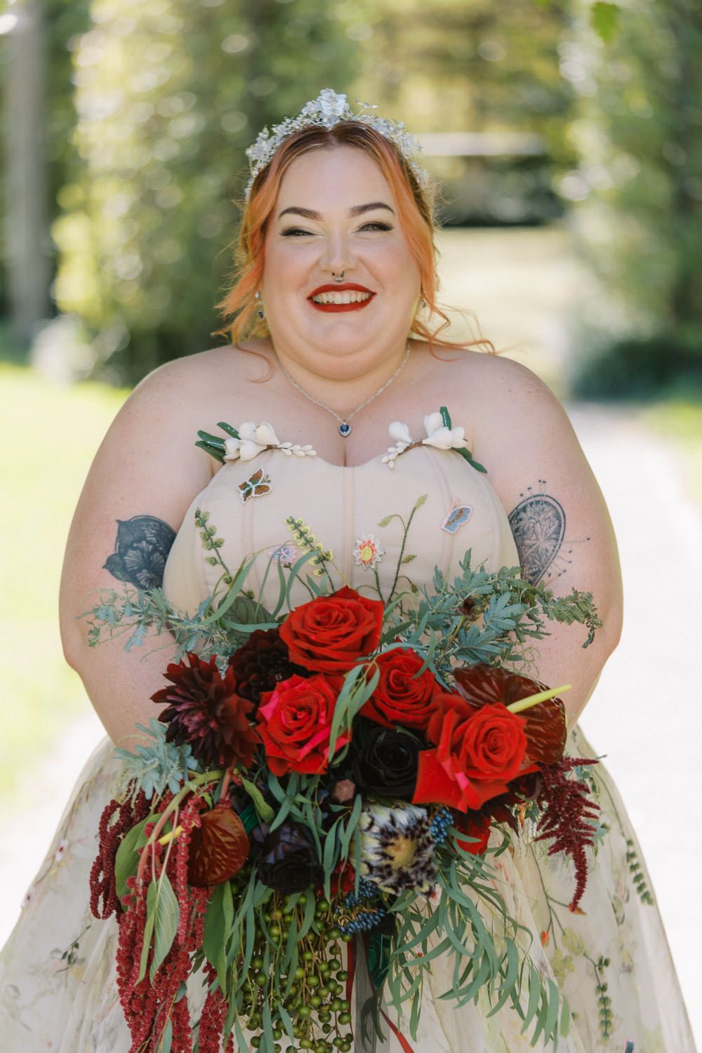 Smiling plus-size bride in floral gown holding red rose bouquet, wearing floral crown and red lipstick outdoors.