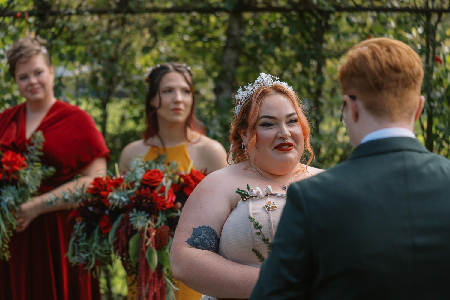 Bride smiling at groom during outdoor wedding ceremony with bridesmaids in red and yellow dresses holding bouquets.