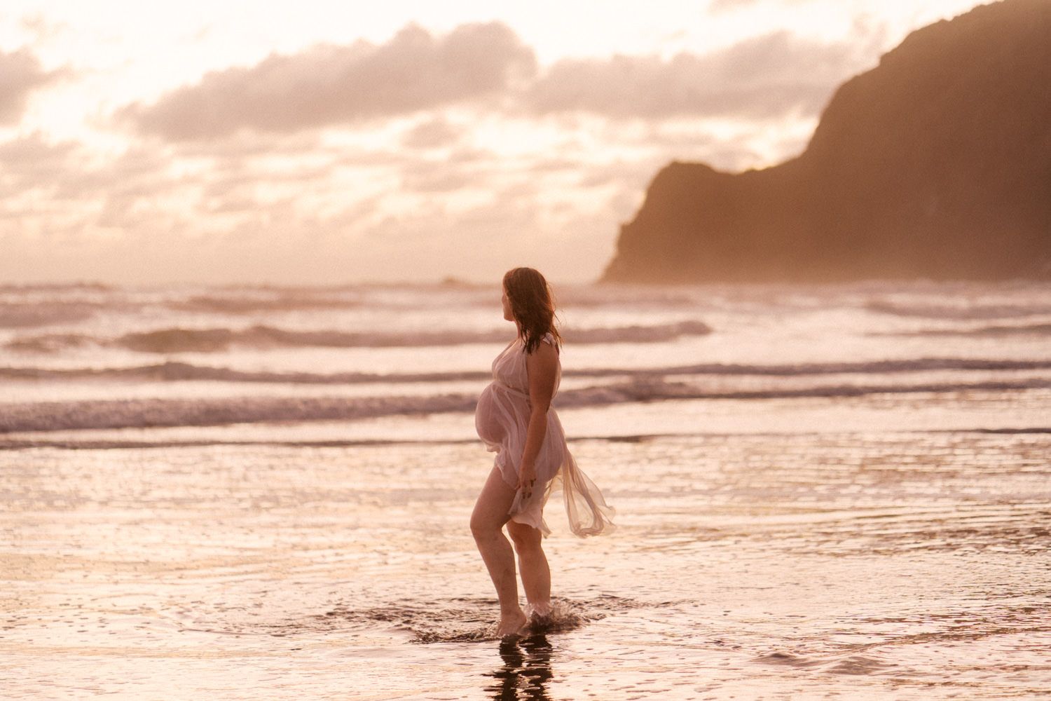 A woman in a white dress walks through ocean waves at sunset on a dramatic beach with misty mountains in the background.