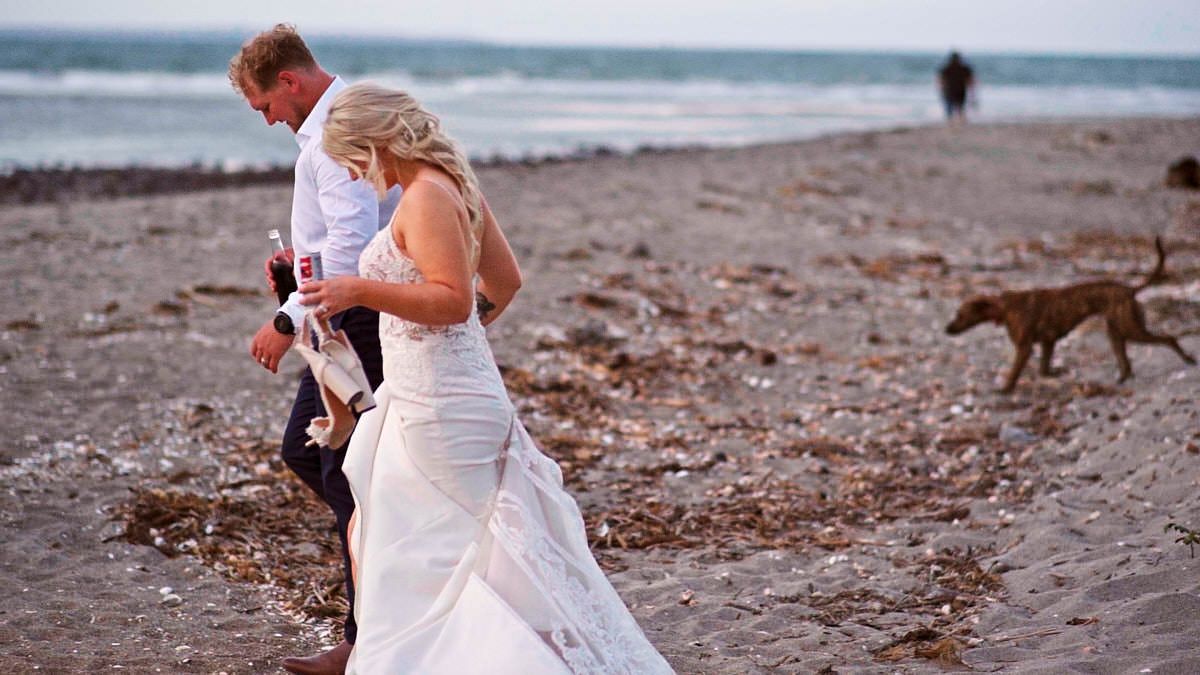 Bride and groom walking on a sandy beach in wedding attire, with a dog and person visible in the background.