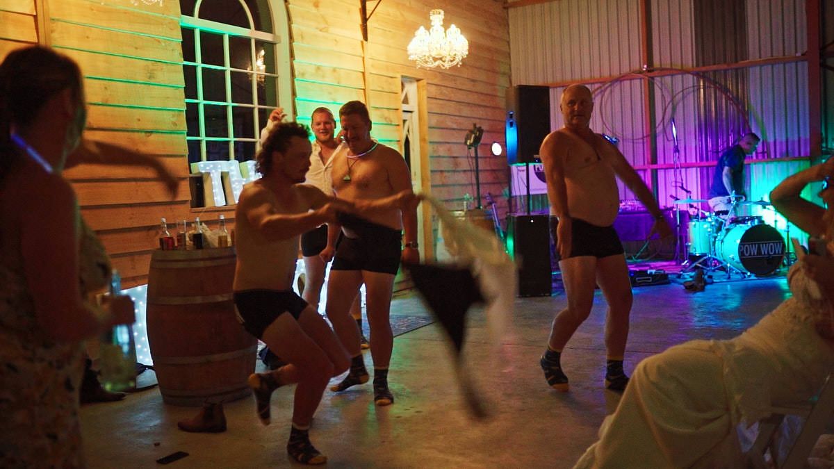 People dancing energetically at an indoor party with live band performing on stage in rustic venue with colorful lighting.