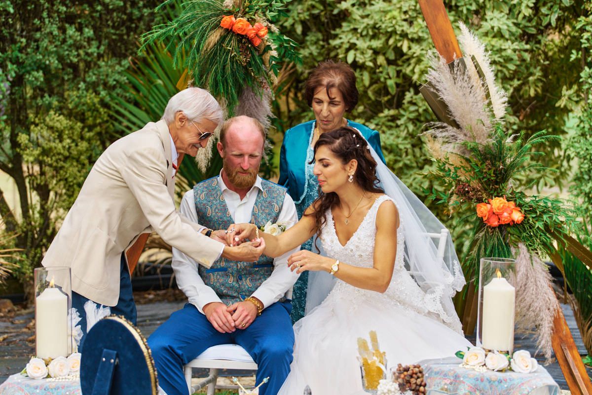 Outdoor wedding ceremony with bride and groom exchanging rings, officiant and witness present, surrounded by lush greenery.