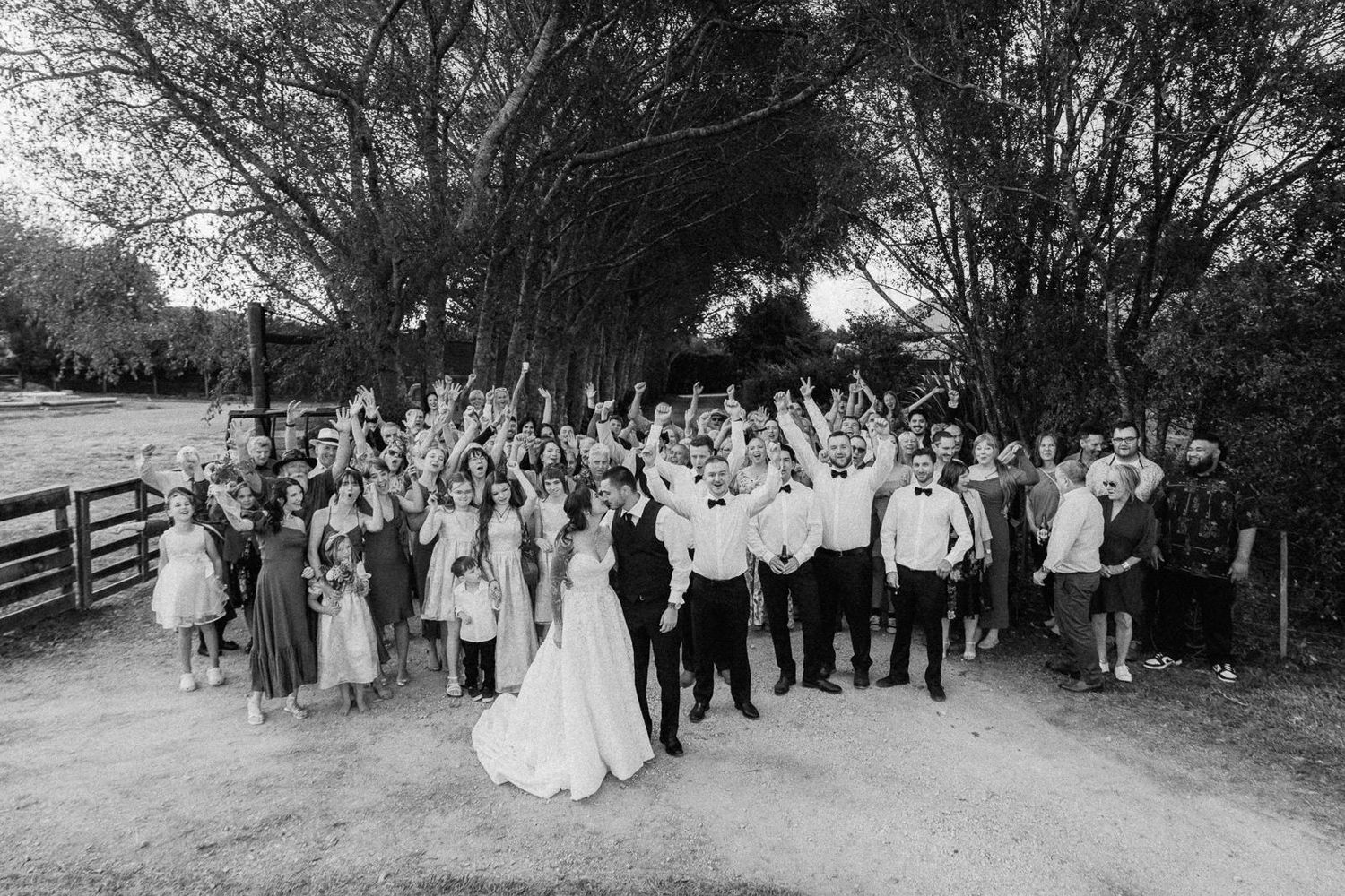Large wedding group photo in black and white with guests cheering outdoors near wooden fence and trees.