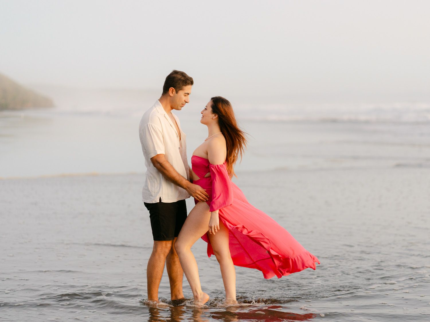 A romantic couple stands in shallow ocean waves, woman in flowing pink dress, man in white shirt and black shorts.