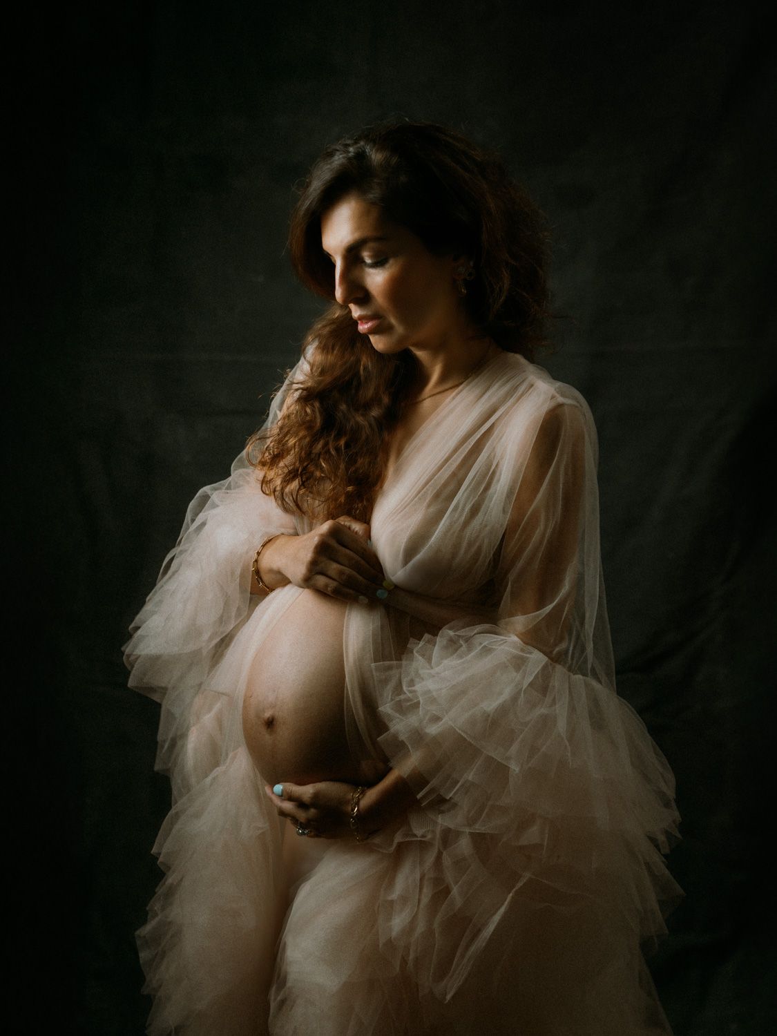 Pregnant woman in sheer white tulle robe cradling her baby bump against a dark studio background.