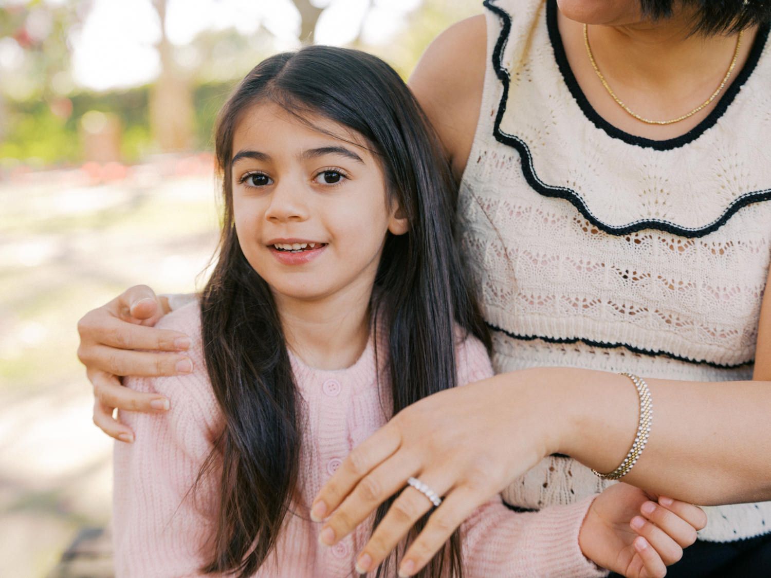 A young girl with long dark hair in a pink top smiles beside a woman in a white lace top outdoors.