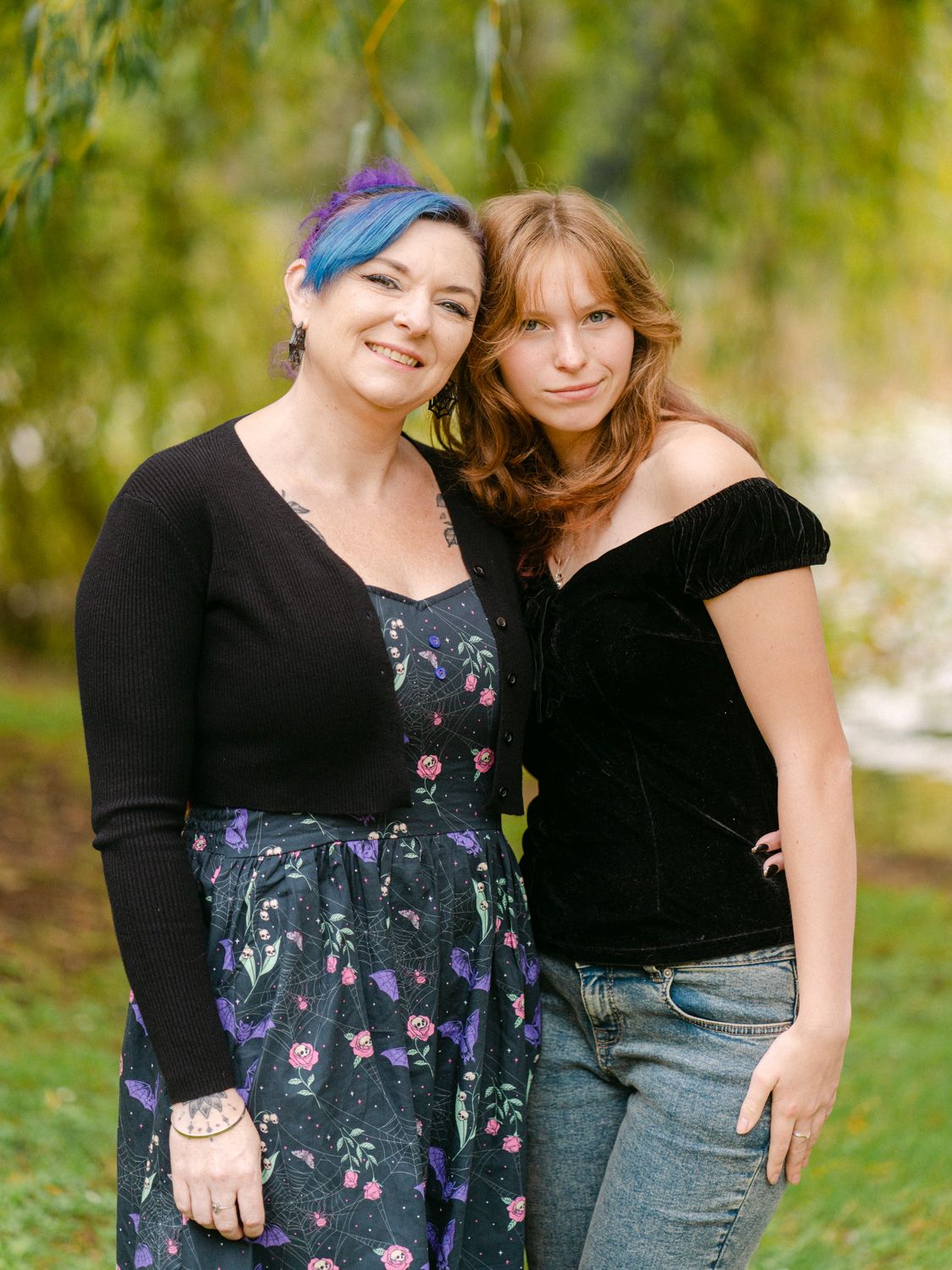 Two women smiling and posing together outdoors in a lush green park setting, one with blue hair wearing a floral dress.