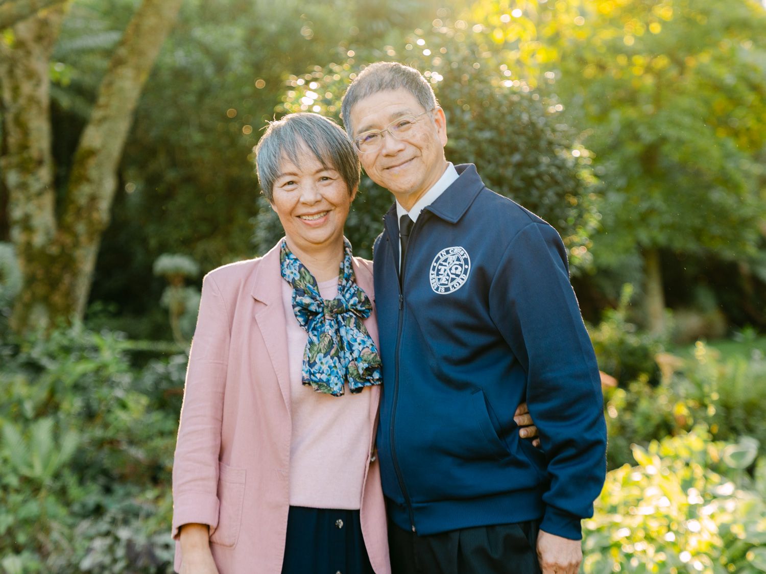 Smiling elderly Asian couple posing together outdoors in a lush green garden setting.