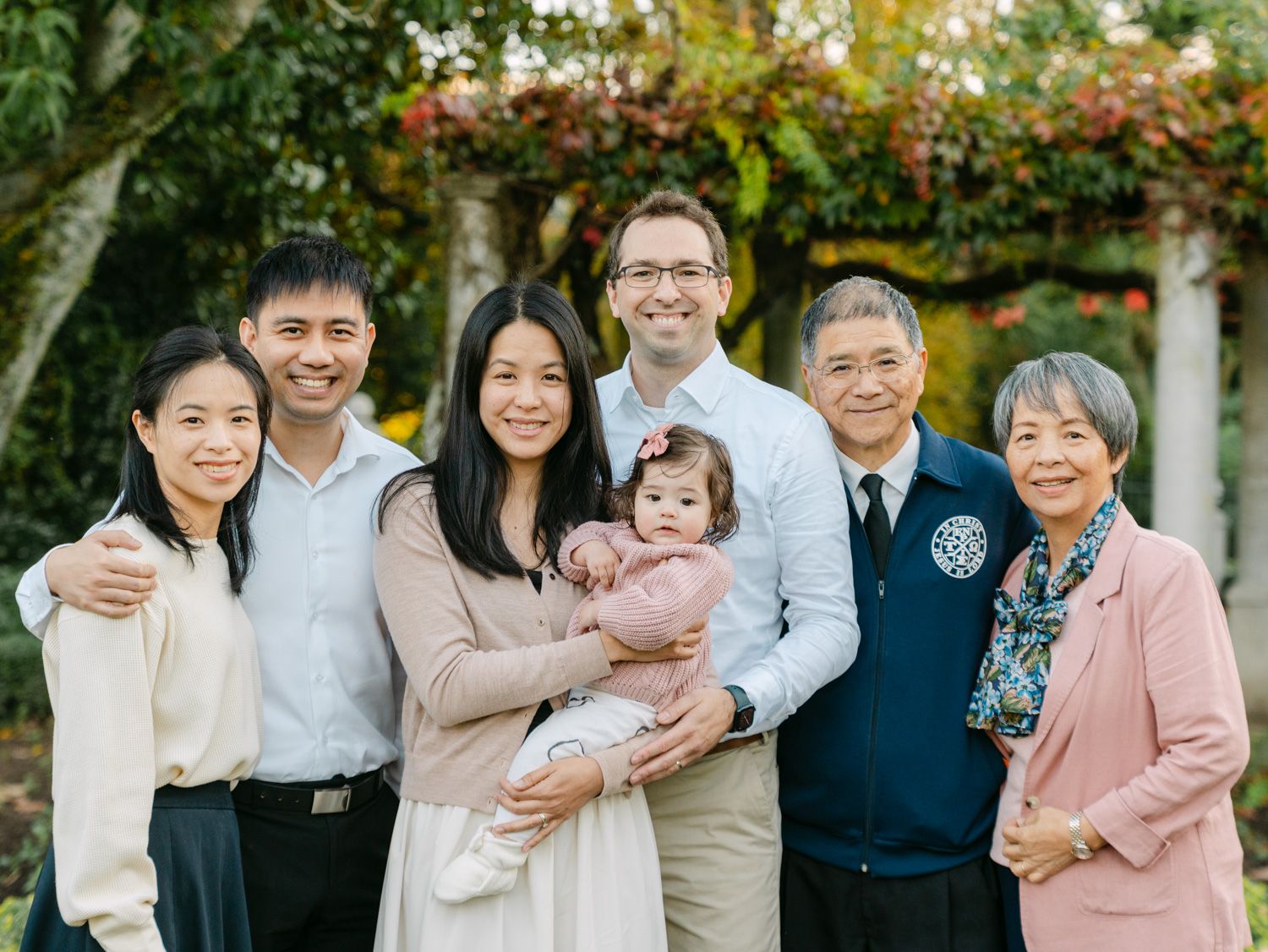 Multi-generational family of seven smiling together outdoors in a lush garden setting for a portrait photo.