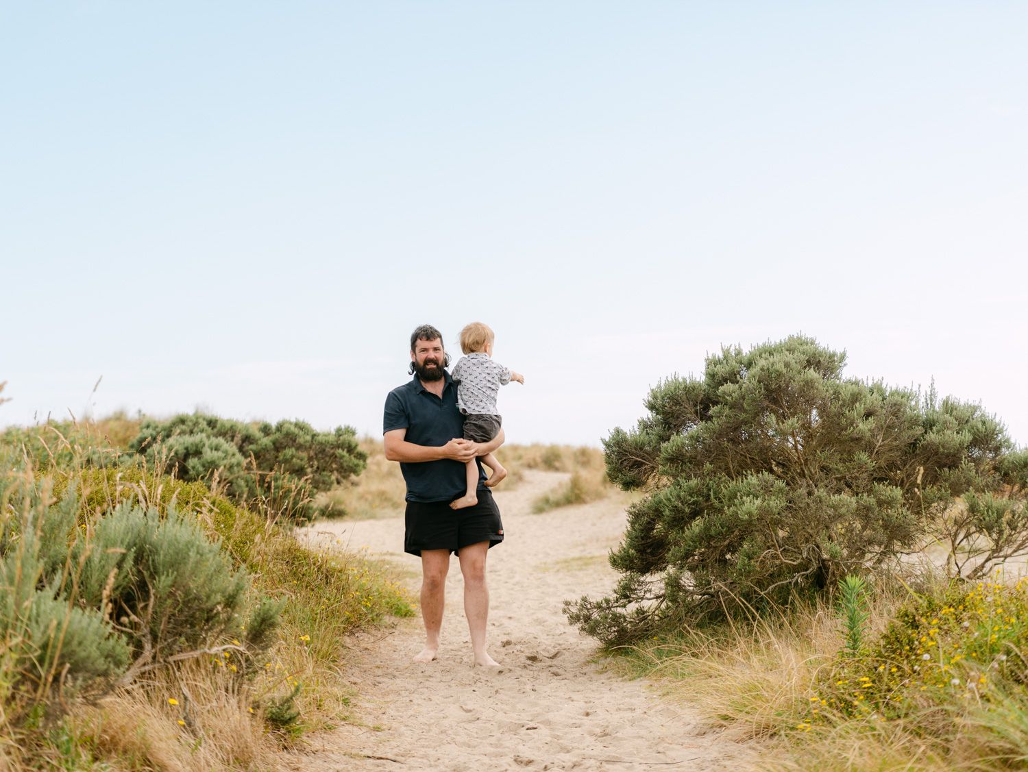 A bearded man holds a toddler while standing on a sandy beach path surrounded by green shrubs on a sunny day.