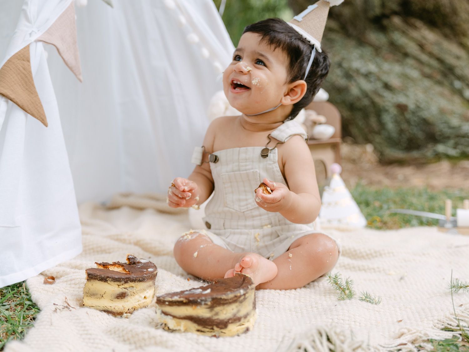 Happy baby in linen romper and party hat smiles while enjoying first birthday cake smash outdoors on blanket.