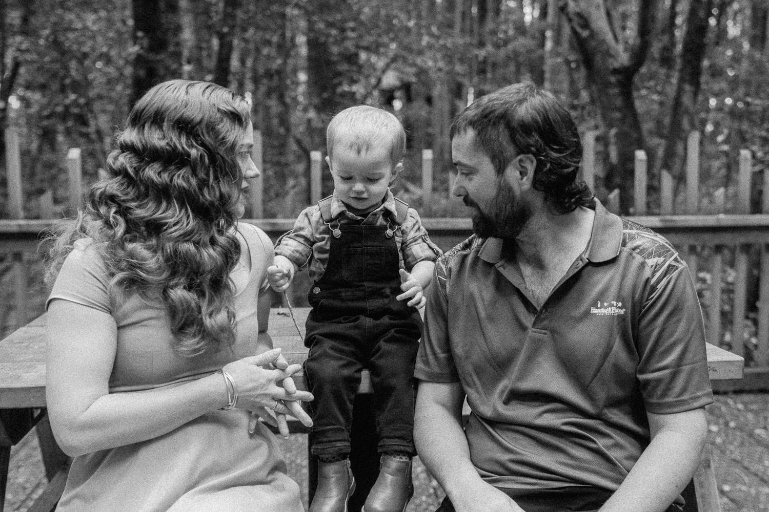 Black and white photo of a young family with a toddler sitting on a bench outdoors in a wooded setting.