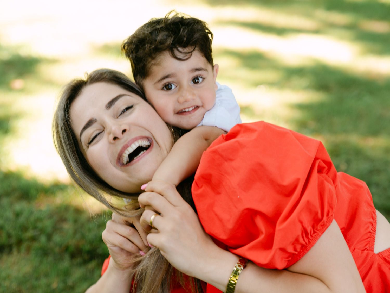 A joyful mother laughing while her young toddler hugs her from behind outdoors on green grass.