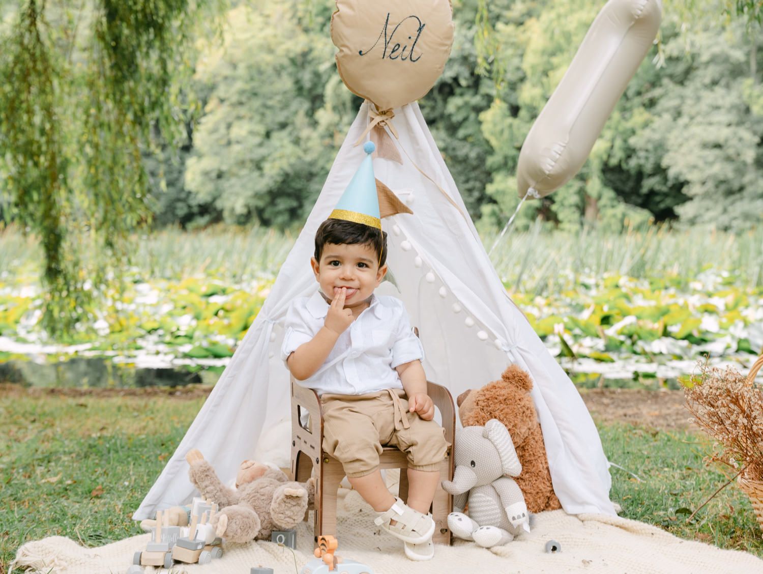 Toddler boy in birthday hat sits before a teepee with stuffed animals and balloons in a lush garden setting.