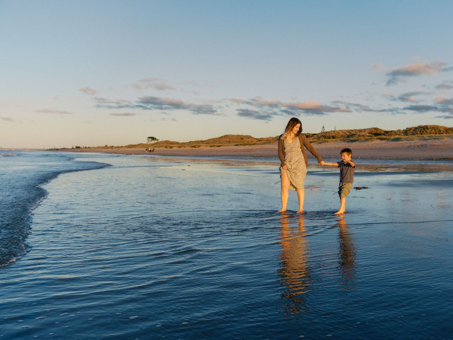 A mother and child hold hands while wading in shallow ocean waves on a sandy beach at golden hour sunset.