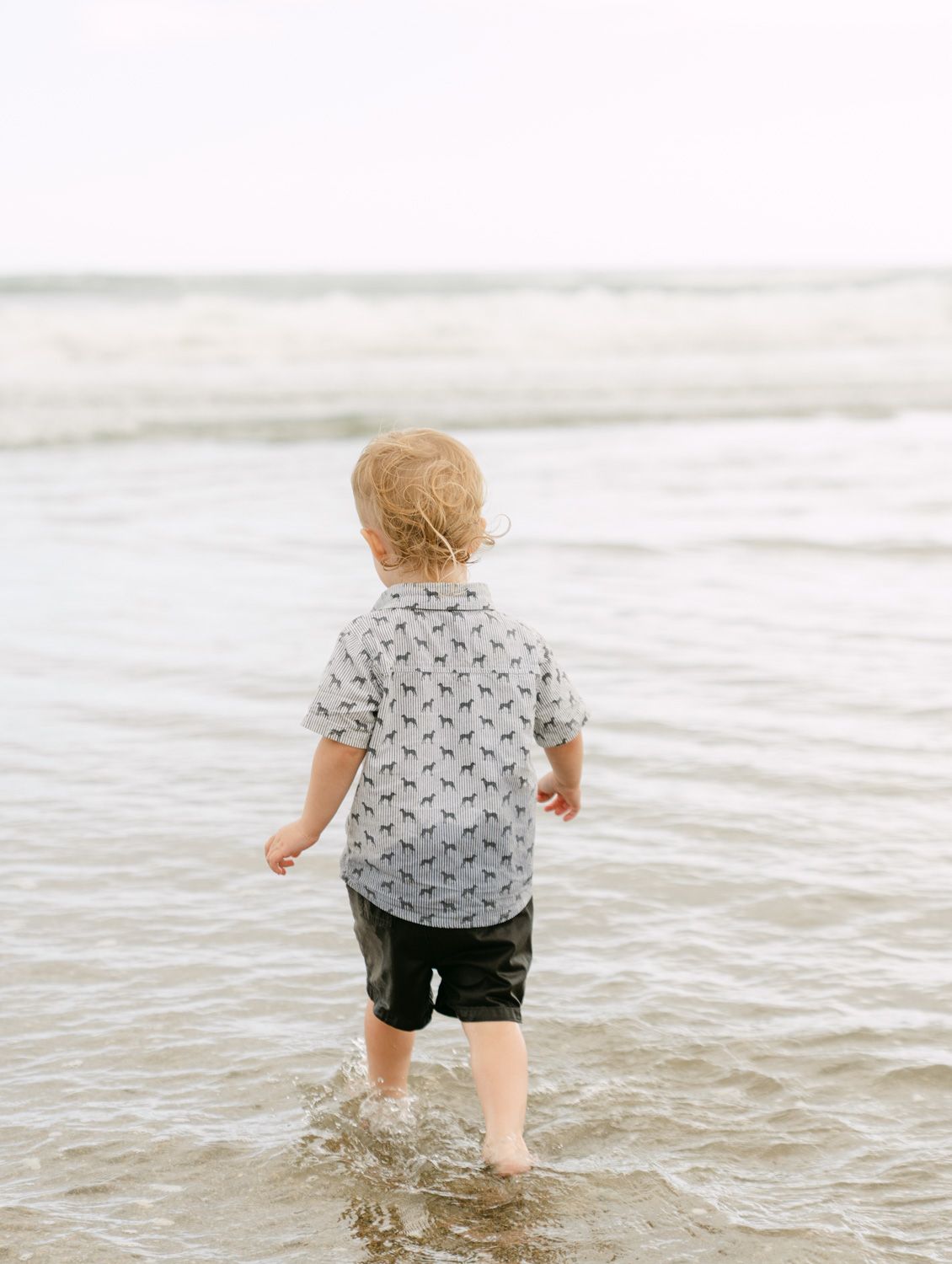 A toddler boy wading in ocean waves at the beach, wearing a patterned shirt and dark shorts, viewed from behind.