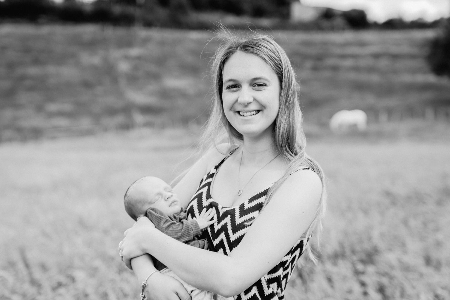 Black and white photo of a smiling young woman holding a newborn baby in an outdoor field setting.