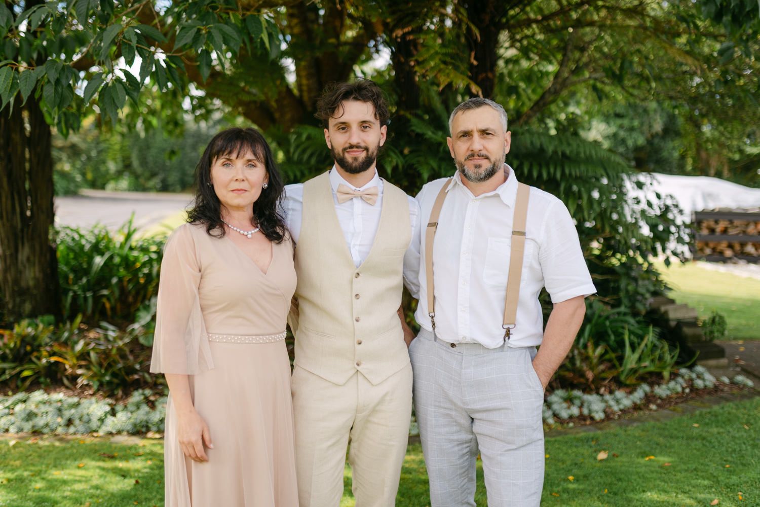 Three elegantly dressed people pose outdoors at a wedding, with lush green trees in the background.