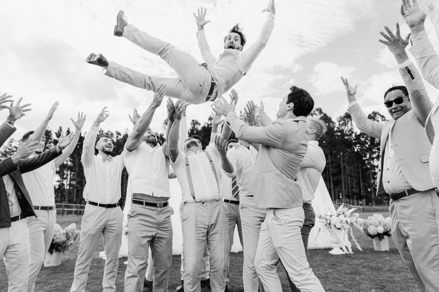 Groomsmen joyfully tossing groom into the air during outdoor wedding celebration in black and white photo.
