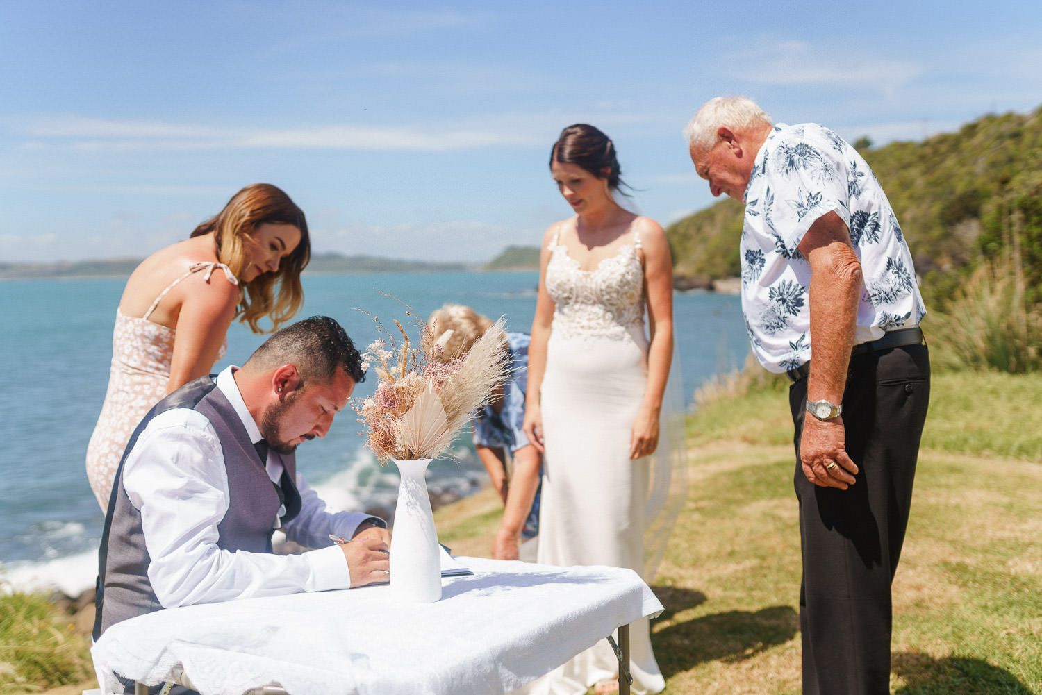 Bride and groom signing wedding documents outdoors by the ocean on a sunny day with guests watching.