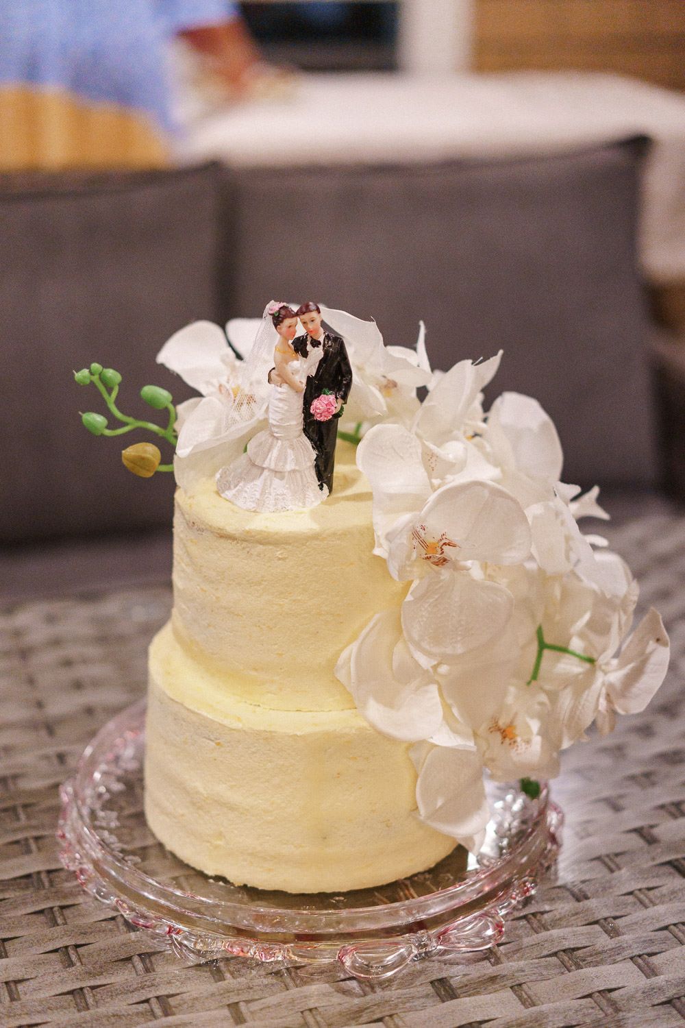 Two-tier white wedding cake with bride and groom topper and white orchids on glass stand.