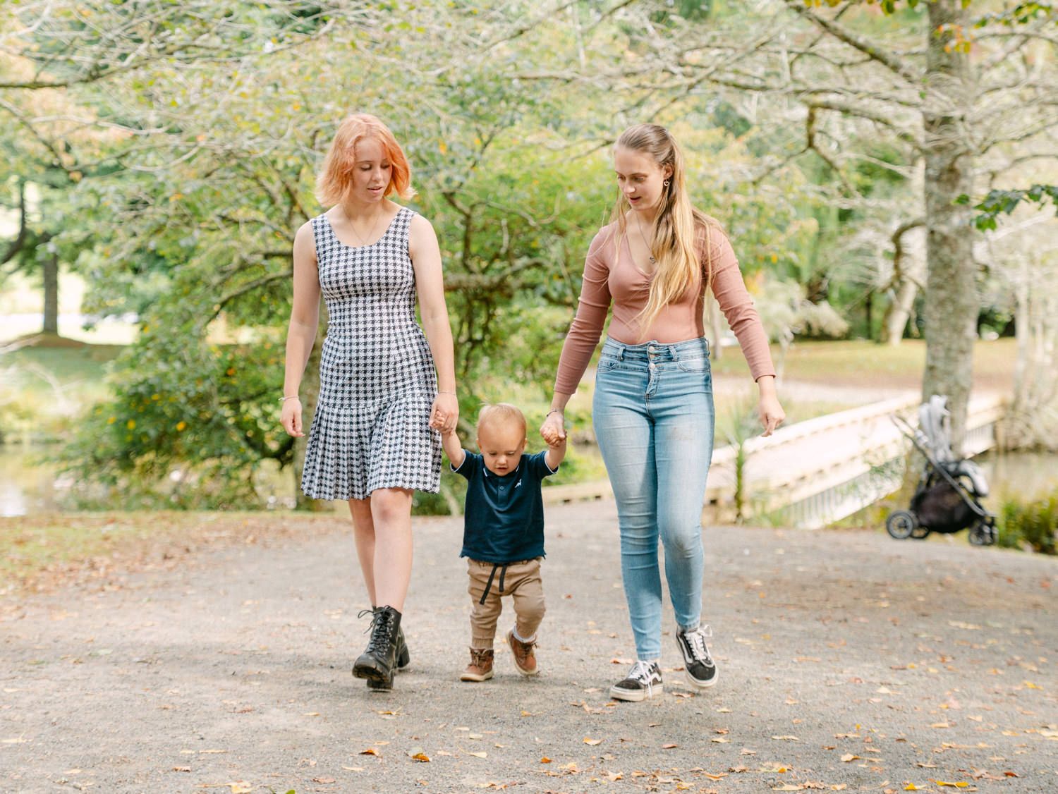 Two women walking with a toddler between them on a park path, holding hands outdoors.