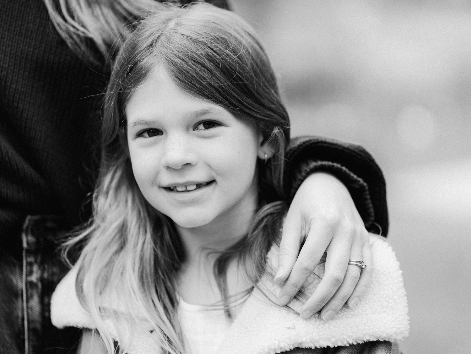Black and white portrait of a smiling young girl with long hair wearing a fleece jacket outdoors.