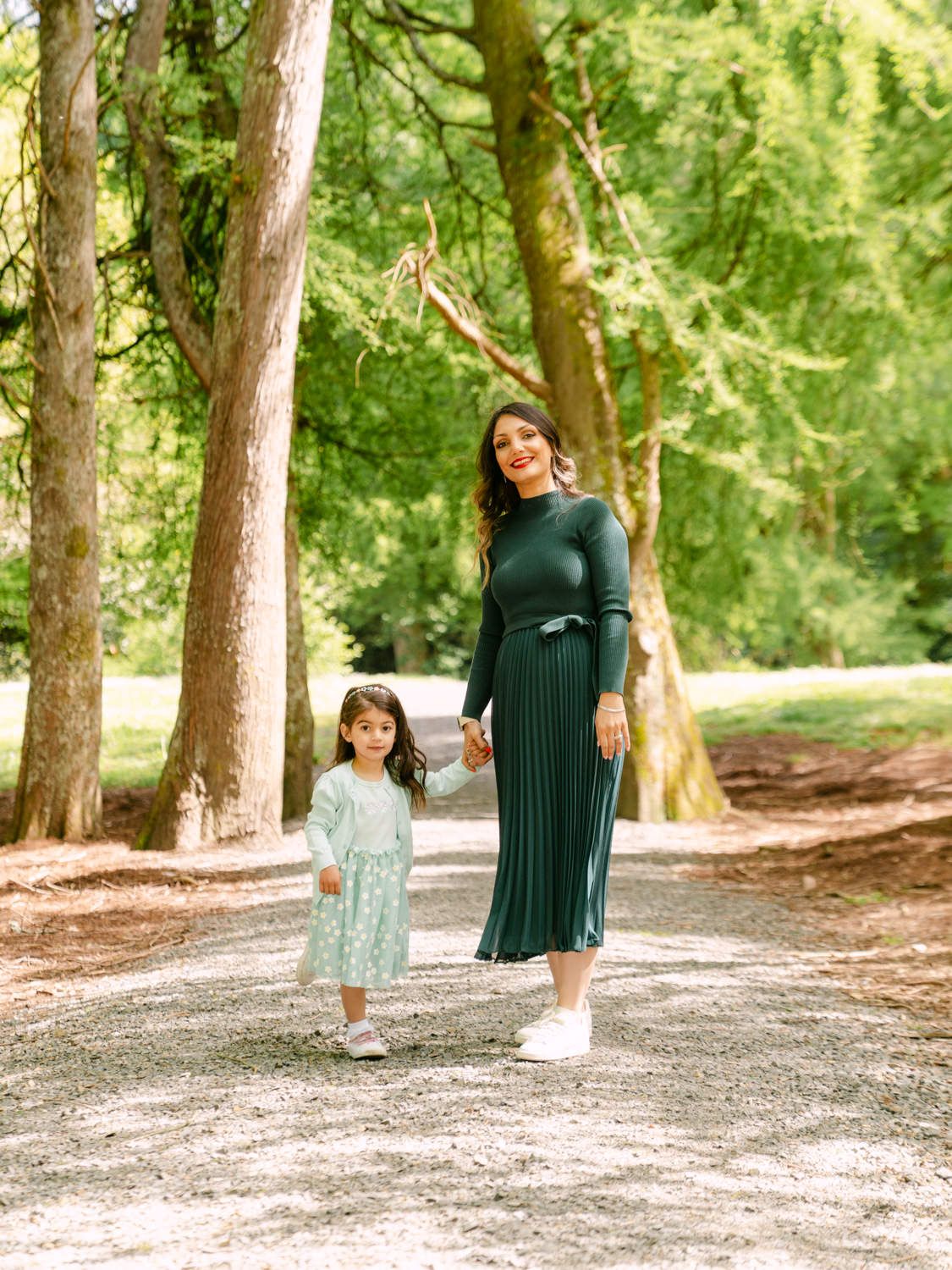 A woman in a green dress holds hands with a young girl in a floral dress while walking on a tree-lined path.